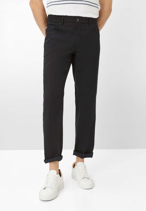 Person iført op rullede sorte bukser og hvide sneakers, stående mod en ensfarvet lys baggrund med hænderne bag ryggen.