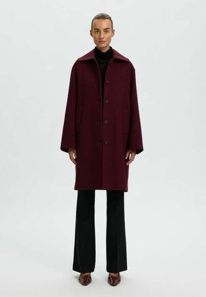 Femme debout portant un manteau bordeaux boutonné sur un haut noir, un pantalon noir et des chaussures pointues bordeaux sur un fond clair uni.