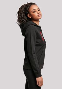 Felpa nera con zip, dotata di una tasca frontale, cappuccio con coulisse, tessuto testurizzato e accento del logo rosso sul petto. Silhouette aderente.