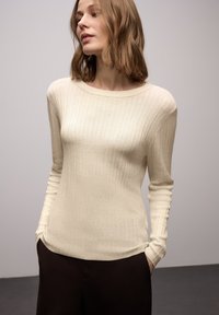 Maglione beige chiaro a coste con scollo a barca e maniche lunghe, caratterizzato da una texture morbida. Abbinato a pantaloni scuri.
