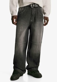 Szerokie czarne dżinsy o subtelnym gradiencie, z wyraźnymi przeszyciami i luźnym krojem, noszone z czarnym paskiem z okrągłymi akcentami metalowymi.