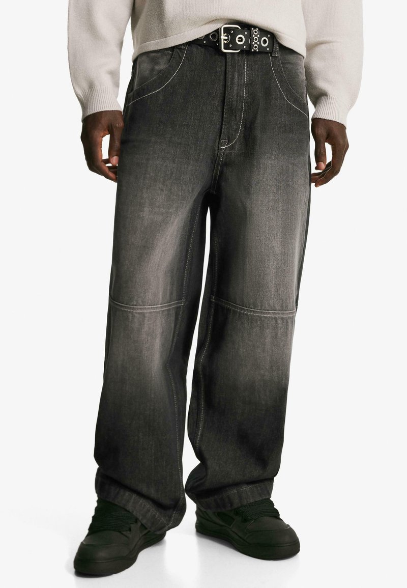 Szerokie czarne dżinsy o subtelnym gradiencie, z wyraźnymi przeszyciami i luźnym krojem, noszone z czarnym paskiem z okrągłymi akcentami metalowymi.