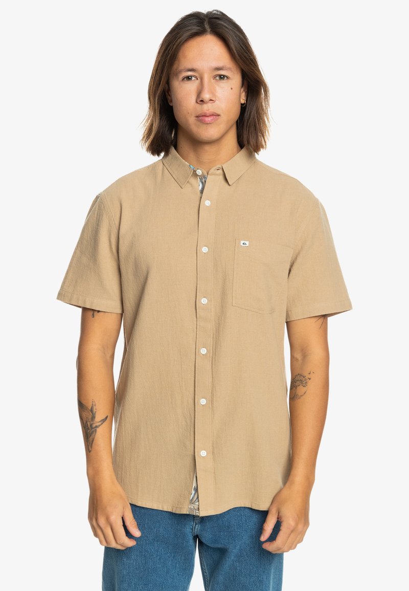 Beige Kurzarmhemd mit Knopfleiste aus strukturiertem Stoff, ausgestattet mit einer Brusttasche und einem gemusterten Futter, das am Saum sichtbar ist.