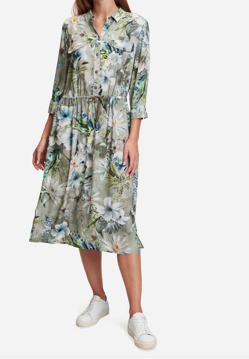 Blommig skjortklänning med trekvartsärmar, cinchad midja och grönfärgad tyg med vita och blå blommor. Kombinerad med vita sneakers.