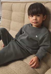 Dunkelgraues Baumwoll-Set aus Pullover und Hose mit rundem Ausschnitt und gerippten Bündchen. Kind sitzt auf einem strukturierten beigen Sofa.