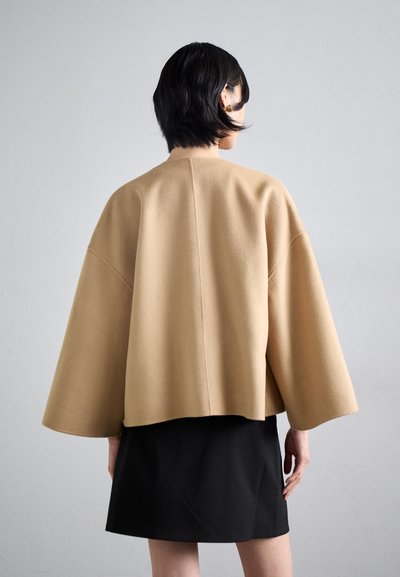 Beige villasegusti lühike jakk ülepaisutatud varrukatega, sirge äärisega ja minimalistliku disainiga. Kanda musta seeliku peal, näidates puhtat siluetti.