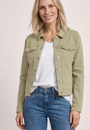 Woman smiling wearing a light olive green buttoned jacket over a white t-shirt and blue jeans against a plain pale background.