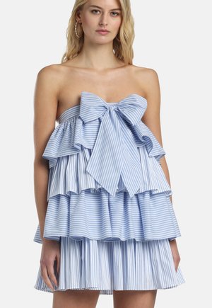 Žena v bezramienkových vrstvených šatách svetlomodrej a bielej pruhovanej farby s veľkou mašľou na hrudi a volánovými vrstvami.