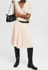 Lyserød plisseret kjole med sort bælte, kombineret med en kortærmet beige top og knæhøje sorte læderstøvler. Sort håndtaske inkluderet.