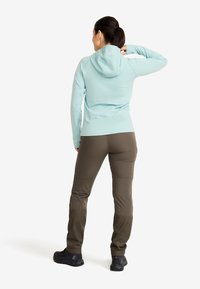 Kvinna står med ryggen vänd mot kameran, iklädd en ljusblå hoodie, bruna friluftsbukser och svarta vandringsskor mot en vit bakgrund.