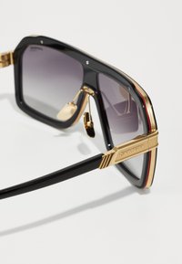 Occhiali da sole neri oversize con lenti sfumate, dettagli in oro sulla montatura e una forma rettangolare elegante caratterizzata da strisce rosse vivaci.