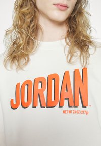 Sweat-shirt blanc avec inscription "JORDAN" en orange vif et accents noirs. Texture douce et col rond. Étiquette de taille visible sous le texte.