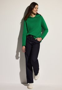 Grön långärmad hoodie med avslappnad passform, kombinerad med mörkblå jeans. Bärs med vita skor, stående mot en ljus bakgrund.
