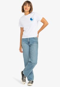 White cotton t-shirt with a blue yin-yang design on the left chest, paired with light blue denim jeans and black sneakers.
