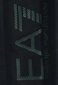 Μαύρο διχτυωτό ύφασμα με λογότυπο σε ανοιχτό πράσινο γεωμετρικό σχέδιο και μερικό κείμενο ορατό μέσα από το μοτίβο του διχτυού.