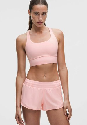 Mujer con sujetador deportivo rosa claro y pantalones cortos a juego, de pie sobre un fondo gris claro liso.