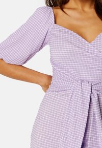 Robe portefeuille à carreaux violets avec des manches bouffantes, décolleté en V et une ceinture à nouer à la taille. Tissu texturé avec des lignes horizontales et verticales.