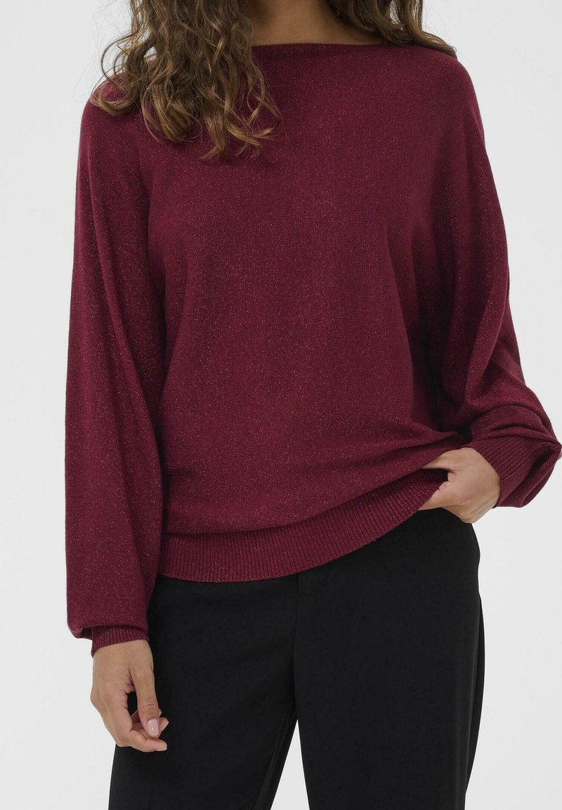 Personne portant un pull bordeaux à manches longues et coupe ample ainsi qu'un pantalon noir, tenant l'ourlet du pull d'une main.