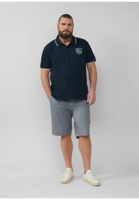 Mann mit Bart, der ein marineblaues Polo-Shirt mit dem Logo "Service Ford Center", graue Shorts und weiße Sneakers trägt, steht vor einem einfarbigen Hintergrund.