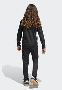 Svart idrottsoverall med långa ärmar och byxor, med vita sidosträck. Skorna inkluderar svarta sneakers med ljus sula.