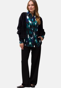 Svart fluffig cardigan över en mönstrad blå-grön skjorta med långa ärmar, tillsammans med lösa svarta byxor och platta svarta skor.