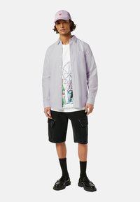 Chemise rayée lilas sur un t-shirt graphique blanc. Shorts cargo noirs, chaussettes noires et chaussures noires brillantes. Casquette avec un cœur rose.
