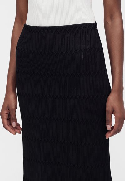 Jupe noire avec un motif texturé vertical portée avec un top blanc côtelé, montrée de la taille jusqu'au milieu de la cuisse, les mains détendues le long du corps.