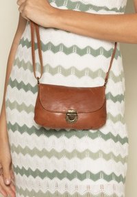 Hnedá kožená crossbody taška s zakriveným tvarom, patentným uzáverom a nastaviteľným popruhom, vystavená na textúrovanom bielom a zeleno vzorovanom pozadí.
