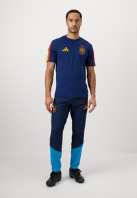 Jūras spēku sporta tērps, kas ietver pieguļošu T-kreklu ar dzelteniem akcentiem un logo, kombinēts ar pieguļošām biksēm jūras zilā un tirkīza krāsā ar svītru detaļām.