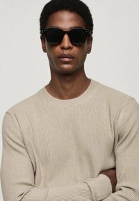 Beige ribbed sweater with a round neckline, featuring long sleeves. Dark, angular sunglasses complete the look, set against a neutral background.
