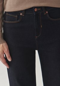 Tamsiai mėlynos džinsinės kelnės su oranžinėmis siūlėmis, mygtuko uždarymu ir daugeliu kišenių. Viršutinė dalis yra smėlio spalvos, su susiūtomis rankovėmis ir apdaila.