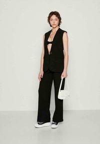 Blazer sem mangas preto sobre uma blusa preta, combinado com calças largas pretas e ténis brancos, segurando uma pequena mala branca.
