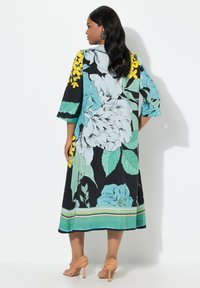 Schwarzes Maxikleid mit floralen Motiven, das große türkisfarbene Blumen und gelbe Akzente zeigt, mit einem gestreiften Saum und entspannten Ärmeln. Glatte Stofftextur.