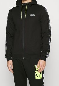Uomo che indossa una felpa nera con zip e il logo EA7 Emporio Armani sul petto e sulle maniche, abbinata a pantaloni neri con un patch del logo EA7 giallo neon.