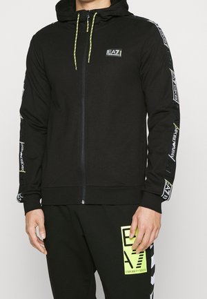 Uomo che indossa una felpa nera con zip e il logo EA7 Emporio Armani sul petto e sulle maniche, abbinata a pantaloni neri con un patch del logo EA7 giallo neon.