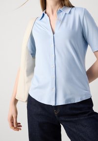 Camicia azzurra a maniche corte con bottoni, realizzata in tessuto morbido, con colletto e orlo curvo. Abbinata a jeans scuri e una borsa chiara.