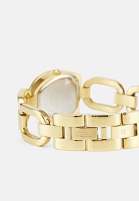 Gold metal wristwatch with a chunky link bracelet and a uniquely shaped case. Features engraved "GUESS" and "G" accents on the clasp.