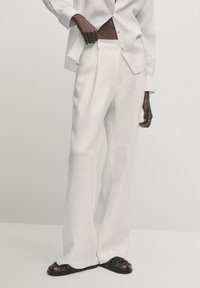 Pantalon large en lin blanc taille haute avec des plis, associé à des sandales noires à brides, sur un fond pâle.