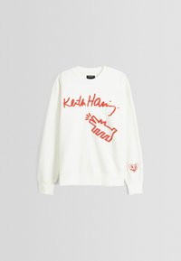 Vit sweatshirt med röd grafisk text "Keith Haring" och en stiliserad handdesign. Inkluderar en röd hjärtlogotyp på ärmen.