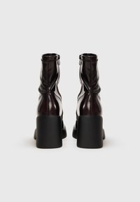 Par de botas tobilleras brillantes de color marrón oscuro con tacones de bloque altos, cremallera trasera y detalles cosidos sobre un fondo claro.