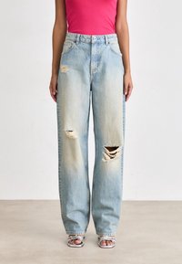 Femme portant un jean large bleu clair déchiré avec de grandes déchirures aux genoux, associé à un haut rose vif et des talons argentés ouverts à l’avant.