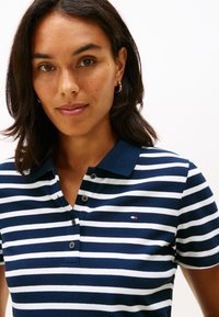 Keskipitkähiuksinen nainen, jolla on kultaiset renkaankorvakorut ja tummansininen, valkoisilla raidoilla varustettu poolopaita, jossa on pieni logo.