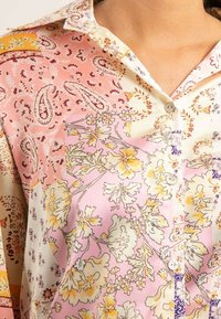 Chemise légère à boutons, ornée d'un mélange de motifs floraux et paisley en rose, jaune et crème, avec des boutons visibles et un col.
