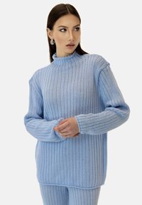 Jasnoniebieski sweter z prążkowanego materiału z wysokim kołnierzem i długimi rękawami. Posiada luźny krój i widoczną teksturę tkaniny.