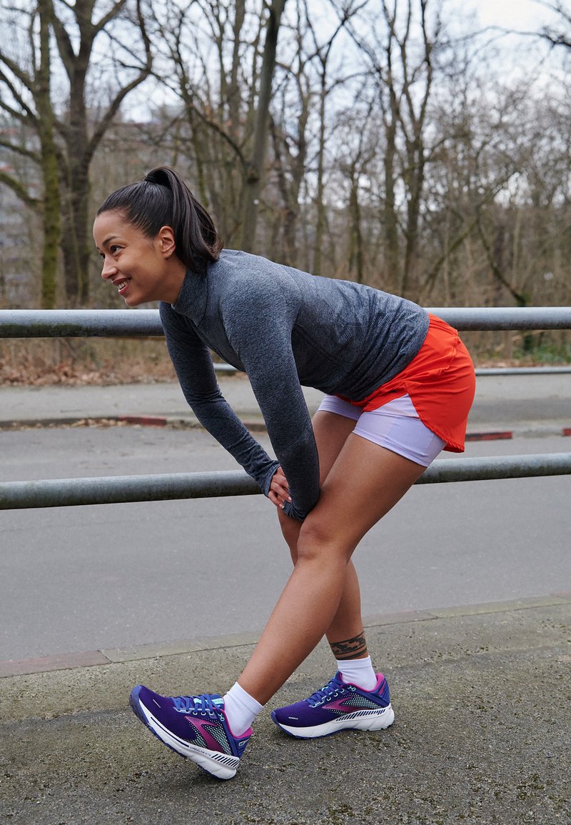 Atleta con una camiseta de manga larga gris y pantalones cortos naranjas, estirándose cerca de una barandilla, vistiendo zapatillas deportivas moradas y blancas. Árboles en el fondo.