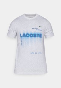 Grå bomullströja med rund halsringning, prydd med texten i blå gradering "LACOSTE" och fraser. Inkluderar grön logga på vänster bröst och blå detaljer.