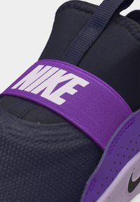 Detailný záber na tmavomodrú a purpurovú tenisku Nike s purpurovým elastickým pásikom, na ktorom je biely nápis Nike.