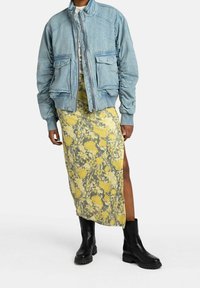 Giacca bomber azzurro chiaro con zip e tasche, abbinata a un lungo vestito floreale giallo con uno spacco laterale e stivaletti neri.