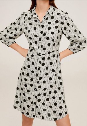 Kobieta w białej sukience na guziki w czarne kropki z trój-ćwiartkowymi bufiastymi rękawami, z rękami opartymi na biodrach.