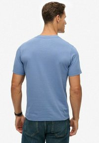 Hombre con una camiseta de manga corta azul claro y pantalones vaqueros azules, visto desde atrás contra un fondo blanco liso.
